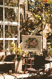 A sunlit garden area features several potted plants and a framed painting on an easel depicting pink flowers. The scene is set against a backdrop of a glass window with vine-covered walls. A foldable chair is placed in the foreground, and various gardening tools are scattered around.