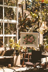A sunlit garden area features several potted plants and a framed painting on an easel depicting pink flowers. The scene is set against a backdrop of a glass window with vine-covered walls. A foldable chair is placed in the foreground, and various gardening tools are scattered around.