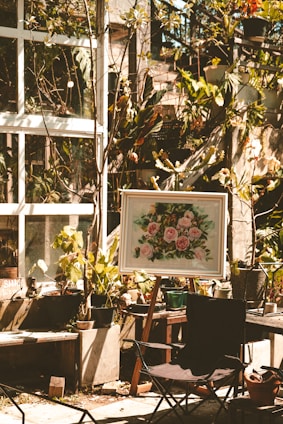 A sunlit garden area features several potted plants and a framed painting on an easel depicting pink flowers. The scene is set against a backdrop of a glass window with vine-covered walls. A foldable chair is placed in the foreground, and various gardening tools are scattered around.