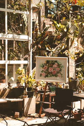 A sunlit garden area features several potted plants and a framed painting on an easel depicting pink flowers. The scene is set against a backdrop of a glass window with vine-covered walls. A foldable chair is placed in the foreground, and various gardening tools are scattered around.