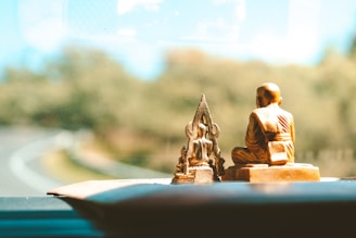 Two small statues, one of a monk and another of a Buddha, are placed on a dashboard inside a vehicle. The background is slightly blurred, showing a curving road and greenery outside through the window.