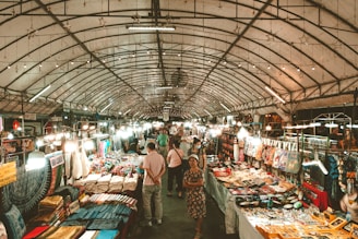 A bustling indoor market filled with people browsing various stalls that display colorful textiles, clothing, and other handcrafted items under a canopy of bright lights.