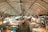 A bustling indoor market filled with people browsing various stalls that display colorful textiles, clothing, and other handcrafted items under a canopy of bright lights.