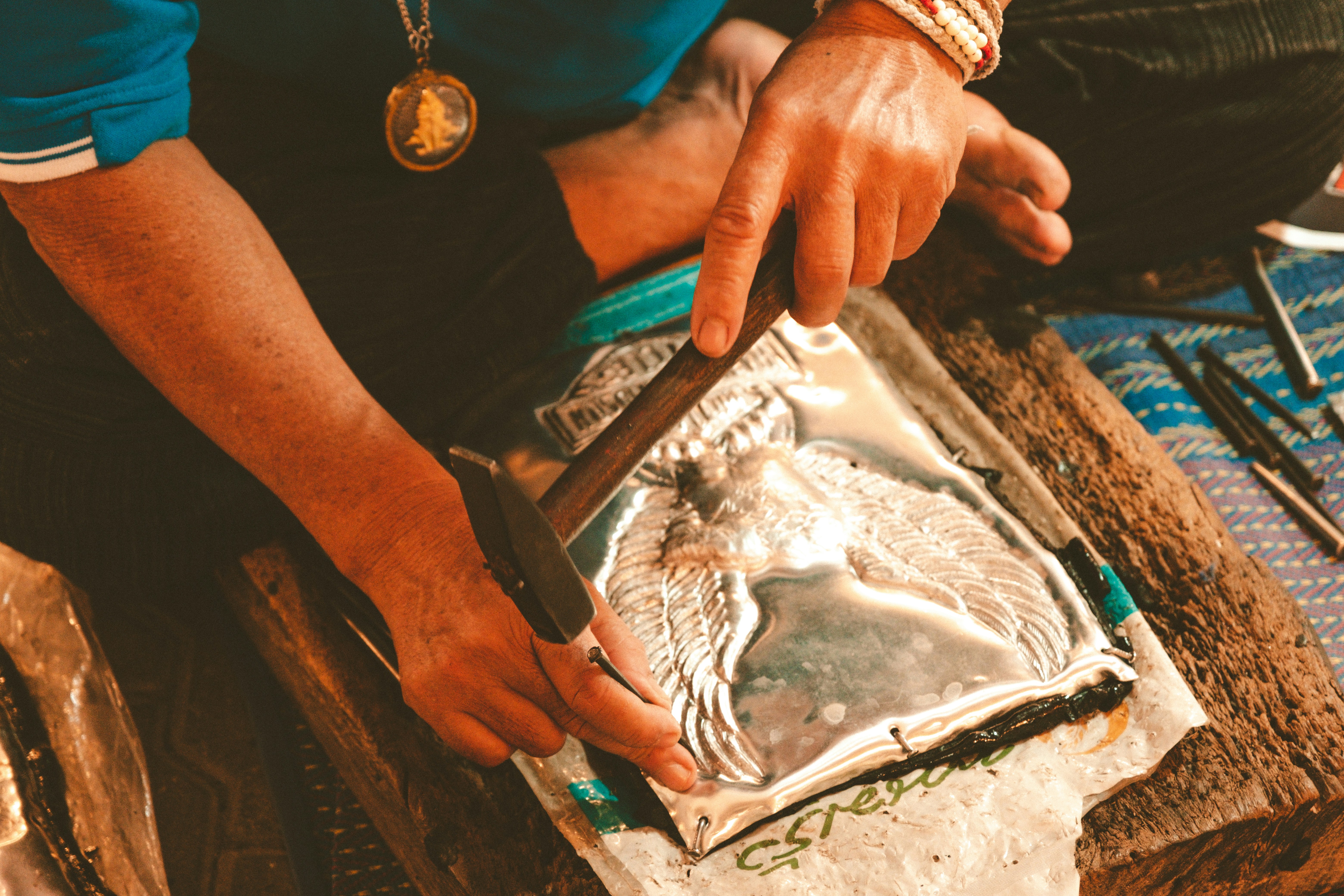 A person is working with metal embossing, using tools to shape a metal sheet with an intricate design. The person is wearing a blue shirt and a necklace with a medallion. The setting appears to be a workshop or craft space.