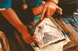 A person is working with metal embossing, using tools to shape a metal sheet with an intricate design. The person is wearing a blue shirt and a necklace with a medallion. The setting appears to be a workshop or craft space.