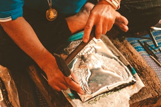 A person is working with metal embossing, using tools to shape a metal sheet with an intricate design. The person is wearing a blue shirt and a necklace with a medallion. The setting appears to be a workshop or craft space.