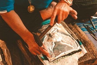 A person is working with metal embossing, using tools to shape a metal sheet with an intricate design. The person is wearing a blue shirt and a necklace with a medallion. The setting appears to be a workshop or craft space.