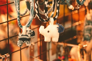 Two fabric animal keychains hang from a display rack. One is a white elephant with a blue tail, while the other is a smaller creature with a blue and white pattern. The background is warmly lit, with a blurred, golden bokeh effect.