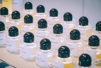 A collection of neatly arranged perfume bottles with black caps and white labels displaying various fragrances. The background is softly blurred, emphasizing the bottles' glass and liquid contents.