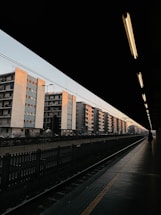 A row of multi-story apartment buildings with a pastel color palette is seen from the perspective of a train station platform. The station roof creates a shadowed frame, leaving the platform and tracks dimly lit. A lone person walks along the platform, and overhead lights cast a warm glow, contrasting with the clear early evening sky.