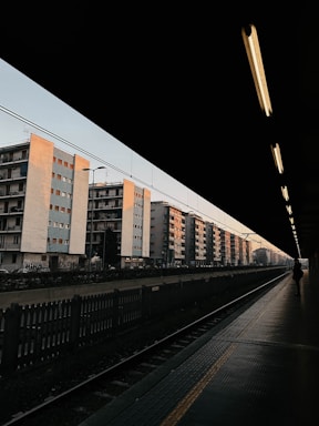 A row of multi-story apartment buildings with a pastel color palette is seen from the perspective of a train station platform. The station roof creates a shadowed frame, leaving the platform and tracks dimly lit. A lone person walks along the platform, and overhead lights cast a warm glow, contrasting with the clear early evening sky.