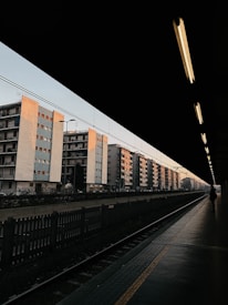 A row of multi-story apartment buildings with a pastel color palette is seen from the perspective of a train station platform. The station roof creates a shadowed frame, leaving the platform and tracks dimly lit. A lone person walks along the platform, and overhead lights cast a warm glow, contrasting with the clear early evening sky.