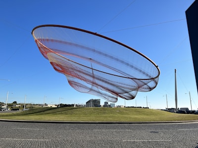 A large, circular net-like structure suspended in the air, supported by multiple cables anchored to the ground. The sculpture appears delicate, with shades of red and white, against a clear blue sky. Below is a grassy hill and a paved road, with street lamps and a few distant buildings visible.