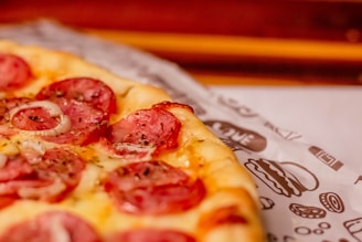 A close-up view of a freshly baked pizza with melted cheese, sliced salami, and thin onion rings on top. The pizza rests on baking paper with printed illustrations of burgers and pizzas.