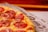 A close-up view of a freshly baked pizza with melted cheese, sliced salami, and thin onion rings on top. The pizza rests on baking paper with printed illustrations of burgers and pizzas.