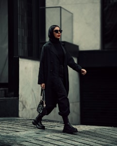 A stylish man confidently wearing a black designer coat and gold-accented accessories in a modern urban setting.