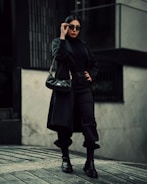 A woman is dressed in an all-black outfit, including a long coat, turtleneck, pants, and boots. She wears sunglasses and carries a black handbag. Her pose is confident and stylish, with one hand on her hip and the other adjusting her sunglasses. The background features an urban setting with monochromatic tones.
