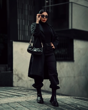 A woman is dressed in an all-black outfit, including a long coat, turtleneck, pants, and boots. She wears sunglasses and carries a black handbag. Her pose is confident and stylish, with one hand on her hip and the other adjusting her sunglasses. The background features an urban setting with monochromatic tones.