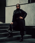 A person wearing a black hijab, long black coat, and sunglasses stands confidently on marble steps. The outfit is accessorized with a black handbag and black boots, creating a bold and stylish appearance. The background is composed of light-colored marble walls, adding contrast to the dark attire.