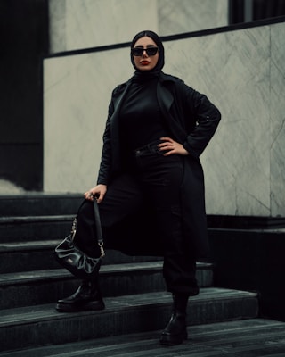 A person wearing a black hijab, long black coat, and sunglasses stands confidently on marble steps. The outfit is accessorized with a black handbag and black boots, creating a bold and stylish appearance. The background is composed of light-colored marble walls, adding contrast to the dark attire.