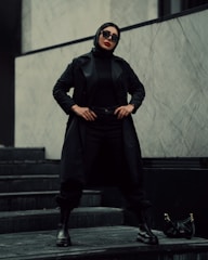 A person stands confidently on a set of dark stairs. Dressed entirely in black, including a long coat, pants, boots, and gloves, they exude a stylish and assertive presence. The individual wears large black sunglasses and a black headscarf, with a bold red lip color. To the side, a small black handbag with a gold chain lies on the step. The background consists of a minimalist, gray marbled wall.