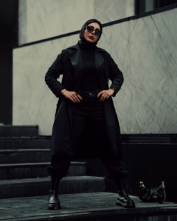A person stands confidently on a set of dark stairs. Dressed entirely in black, including a long coat, pants, boots, and gloves, they exude a stylish and assertive presence. The individual wears large black sunglasses and a black headscarf, with a bold red lip color. To the side, a small black handbag with a gold chain lies on the step. The background consists of a minimalist, gray marbled wall.