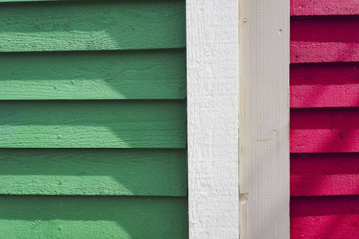 Wooden siding panels are arranged vertically in three distinct colors: green, white, and red. The texture of the wood is visible, with shadow patterns adding depth and contrast.