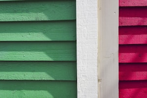 Wooden siding panels are arranged vertically in three distinct colors: green, white, and red. The texture of the wood is visible, with shadow patterns adding depth and contrast.