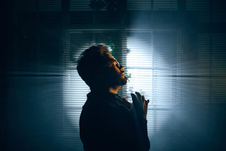 Side profile of Profit Elijah gazing through rain-speckled window, ivory light casting soft shadows on his face.