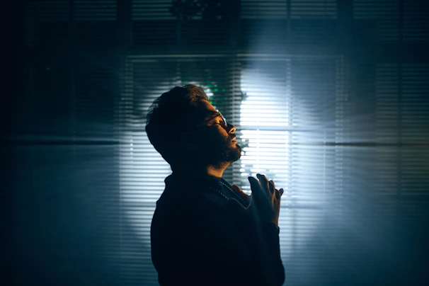 Side profile of Profit Elijah gazing through rain-speckled window, ivory light casting soft shadows on his face.