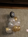 A bouquet of balloons, predominantly in white and gold, is attached to a single black balloon with the number 80 printed in sparkling silver. The balloons are set against a textured, beige wall. The setting suggests a celebration or milestone event, possibly an 80th birthday.