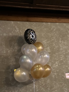 A bouquet of balloons, predominantly in white and gold, is attached to a single black balloon with the number 80 printed in sparkling silver. The balloons are set against a textured, beige wall. The setting suggests a celebration or milestone event, possibly an 80th birthday.
