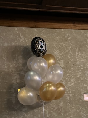 A bouquet of balloons, predominantly in white and gold, is attached to a single black balloon with the number 80 printed in sparkling silver. The balloons are set against a textured, beige wall. The setting suggests a celebration or milestone event, possibly an 80th birthday.