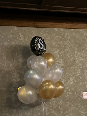 A bouquet of balloons, predominantly in white and gold, is attached to a single black balloon with the number 80 printed in sparkling silver. The balloons are set against a textured, beige wall. The setting suggests a celebration or milestone event, possibly an 80th birthday.