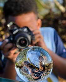 A person is holding a camera and taking a photograph through a transparent glass sphere that reflects the surroundings in a distorted manner. The sphere shows an inverted reflection of the photographer, capturing a mix of greenery and sky in the background.