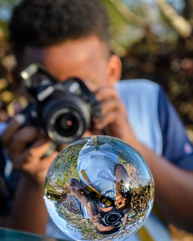 A person is holding a camera and taking a photograph through a transparent glass sphere that reflects the surroundings in a distorted manner. The sphere shows an inverted reflection of the photographer, capturing a mix of greenery and sky in the background.