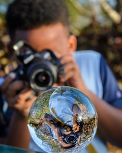 A person is holding a camera and taking a photograph through a transparent glass sphere that reflects the surroundings in a distorted manner. The sphere shows an inverted reflection of the photographer, capturing a mix of greenery and sky in the background.