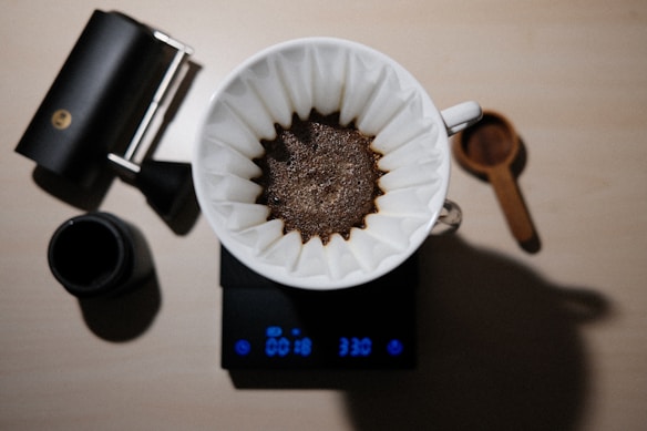 A top-down view of a coffee brewing setup with a white filter containing freshly brewed coffee placed over a black electronic scale. Nearby, there's a black cylindrical grinder, a black mug, and a brown wooden scoop on a light beige surface.