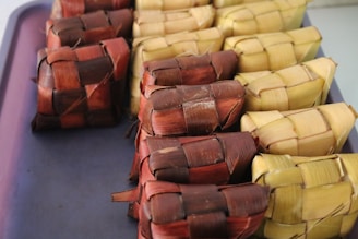 A collection of woven pouches, traditionally known as ketupat, made from coconut or palm leaves, arranged neatly on a tray. The pouches display two primary colors: brown and light yellow, and are tightly packed in a grid-like pattern.