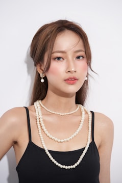 A woman with long brown hair and natural makeup wearing a black top and pearl jewelry, including earrings and a multi-strand necklace. The background is plain white, providing a clean and minimalist look.