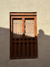 A traditional wooden window with an intricate design is set in a textured beige wall. The window has rectangular panels with circular metal studs and decorative cutouts at the bottom. Light and shadow play across the surface, casting a soft silhouette on the right side.