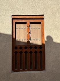 A traditional wooden window with an intricate design is set in a textured beige wall. The window has rectangular panels with circular metal studs and decorative cutouts at the bottom. Light and shadow play across the surface, casting a soft silhouette on the right side.