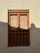 A traditional wooden window with an intricate design is set in a textured beige wall. The window has rectangular panels with circular metal studs and decorative cutouts at the bottom. Light and shadow play across the surface, casting a soft silhouette on the right side.