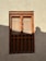 A traditional wooden window with an intricate design is set in a textured beige wall. The window has rectangular panels with circular metal studs and decorative cutouts at the bottom. Light and shadow play across the surface, casting a soft silhouette on the right side.