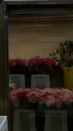 A selection of pink and red flowers wrapped in plastic is displayed on white stands inside a glass cooler, with leafy green plants in yellow containers visible to the right.