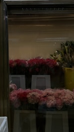 A selection of pink and red flowers wrapped in plastic is displayed on white stands inside a glass cooler, with leafy green plants in yellow containers visible to the right.