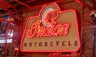 A vintage neon sign with the words 'Indian Motorcycle' prominently displayed, featuring a classic design with a red, glowing border. The background consists of an exposed brick wall, creating an industrial aesthetic. A partial view of a motorcycle and shelving are visible to the left.