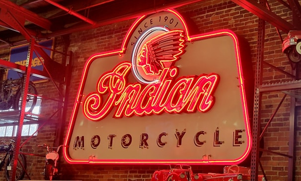 A vintage neon sign with the words 'Indian Motorcycle' prominently displayed, featuring a classic design with a red, glowing border. The background consists of an exposed brick wall, creating an industrial aesthetic. A partial view of a motorcycle and shelving are visible to the left.