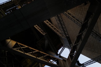 An intricate view of architectural structures featuring a complex network of steel beams and rivets. The image focuses on the underside of a large bridge or similar construction with overlapping geometric patterns.