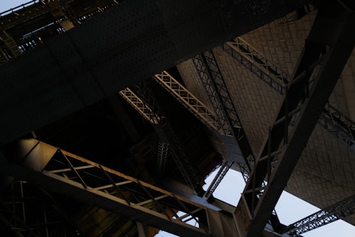 An intricate view of architectural structures featuring a complex network of steel beams and rivets. The image focuses on the underside of a large bridge or similar construction with overlapping geometric patterns.